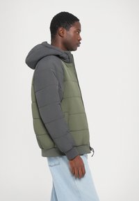 Padded bunda so sivou kapucňou a rukávmi, zeleným telom, rebrovanými manžetami a zipsovým zapínaním. Obsahuje prešívané časti pozdĺž strán.