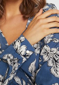 Kvinna i en blå blommönstrad blus med guldknappar, vänster hand vilar på höger axel, visar nyckelbenet och hårstrån.