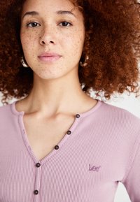Haut Henley côtelé rose clair avec accents de boutons et logo brodé sur la poitrine gauche. Tissu texturé et design en V.