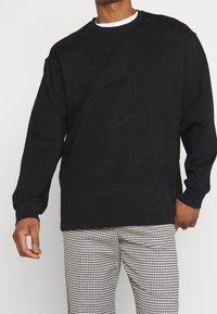 Sweatshirt noir à manches longues avec col rond, poignets et ourlet côtelés. Superposé sur une chemise blanche. Associé à un pantalon à motif pied-de-poule.