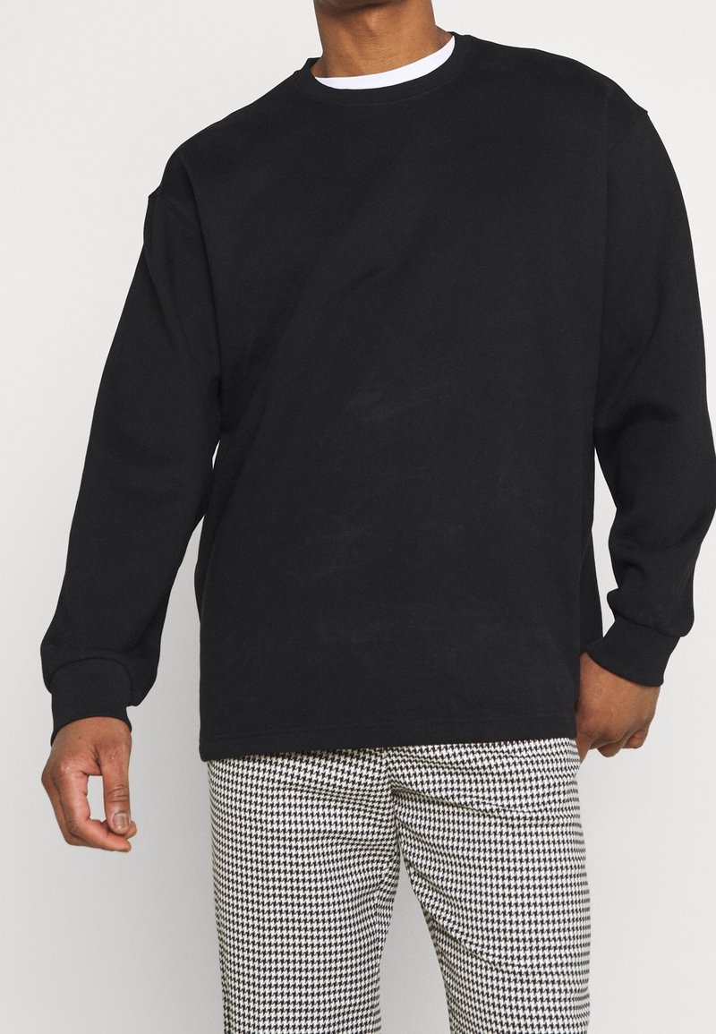 Sweatshirt noir à manches longues avec col rond, poignets et ourlet côtelés. Superposé sur une chemise blanche. Associé à un pantalon à motif pied-de-poule.