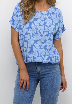 Femme portant un chemisier bleu à manches courtes à motif floral, rentré dans un jean bleu taille haute, debout devant un fond uni.