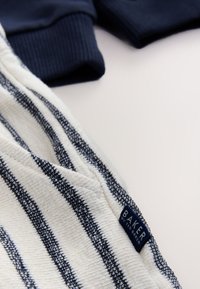 Gestreepte stof in off-white en marineblauw met een gestructureerd oppervlak. Bevat een blauw label met de tekst "BAKER by Ted Baker" vlak bij de naad.