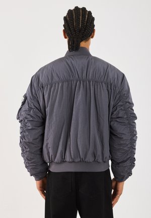 Personne aux cheveux tressés attachés en arrière portant une veste bomber ample, grise et froncée, debout, de dos.