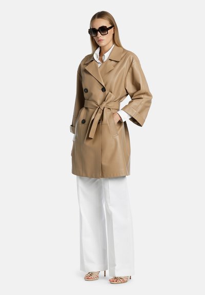 Beige kunstlæder trench coat med dobbeltradet design, store knapper, bælte og brede ærmer, parret med hvide bukser.