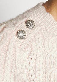 Jasnoróżowy sweter z dzianiny z wzorem w warkocze oraz dwoma okrągłymi guzikami w srebrnym odcieniu ozdobionymi przezroczystymi cyrkoniami.