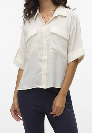 Vrouw draagt een losse witte blouse met knopen en opgerolde mouwen en een donkere broek, geaccentueerd met gelaagde gouden kettingkettingen.