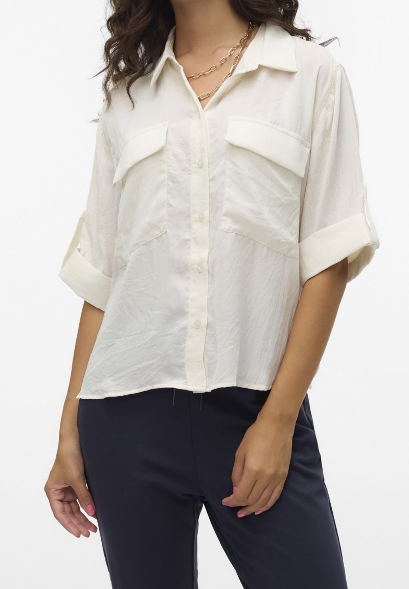 Femme portant une chemise blanche ample boutonnée avec manches retroussées et un pantalon sombre, accessoirisée de colliers chaînes dorés superposés.