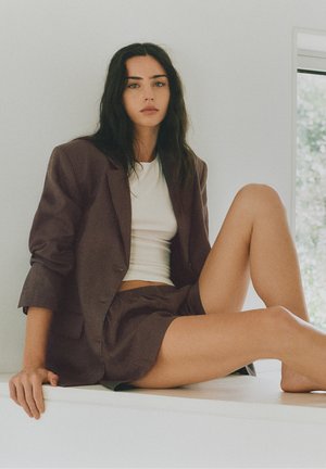 Femme aux longs cheveux foncés portant une veste de costume marron et un short, ainsi qu'un crop top blanc, assise avec une jambe repliée sur une surface blanche.