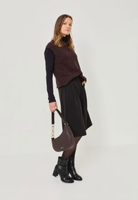 Un haut en tricot sans manches marron sur une chemise à manches longues noire, associé à un short noir plissé, des bottes à talons noires et un sac à épaule marron.