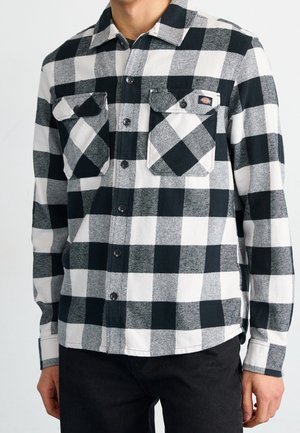 Homme portant une chemise à carreaux noir et blanc à boutons avec poches poitrine et un pantalon noir sur fond blanc.