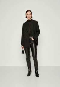 Sacou negru cu dungi fine peste un pulover negru, asortat cu pantaloni din piele neagră strălucitoare și ghete până la gleznă. Modelul ține o geantă mică.