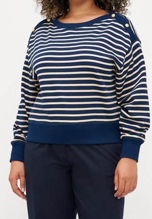Person wearing navy blue and beige striped long-sleeve top with gold buttons on shoulders and navy blue pants, shown from neck to hips.
