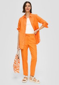 Orangenes Leinenhemd mit hochgerollten Ärmeln, passenden Jogginghosen mit Kordelzug und einem weißen Tanktop. Dazu gehören beiges Sandalen und eine gestreifte Tragetasche.