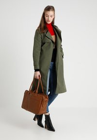Trench coat verde oliva, dolcevita nera e jeans blu. Borsa marrone con due manici. Stivaletti neri con tacco. Sfondo semplice.