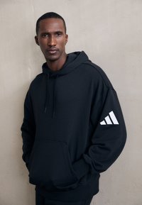 Mann trägt einen schwarzen Hoodie mit einem weißen Adidas-Logo auf dem linken Ärmel und steht vor einer strukturierten beigefarbenen Wand.