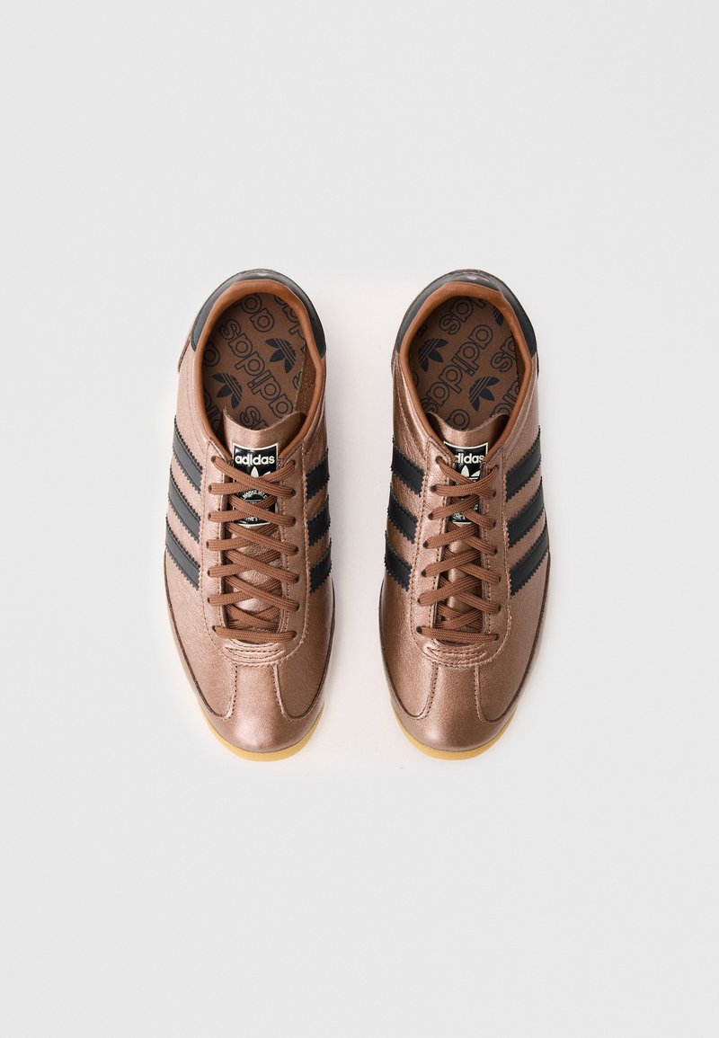 Sneaker in pelle marrone con design a tre strisce nere, chiusura con lacci, suola in gomma e logo Adidas impresso sulla linguetta e sulla soletta.