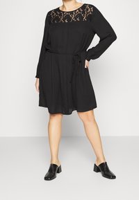 Svart klänning med långa ärmar, spetstopp, åtsittande midja och knälång kjol. Bärs med svarta klackmules. Slät tygtextur.