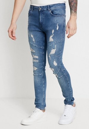 Slim-fit blue jeans with distressed patches and paint splatters, worn with white sneakers and a white shirt on a standing person.