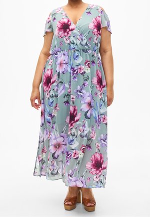 Femme portant une robe longue à fleurs vert clair avec des fleurs roses et violettes, des manches à épaules dénudées et des sandales compensées marron.