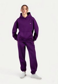 Paarse trui met capuchon en voorzak, gecombineerd met bijpassende broek. Beide kledingstukken hebben een losse pasvorm en een zachte textuur.