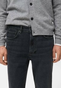 Cardigan gris con botones negros sobre jeans oscuros. Los jeans tienen un corte clásico con bolsillos frontales y una sutil textura desvaída.