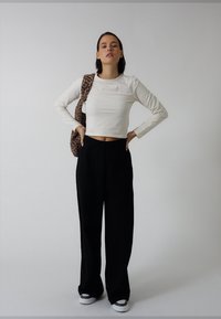 Witte crop top met lange mouwen, zwarte wijde broek en patroonrijke bruine tas. Model draagt grijze sneakers. Minimalistische achtergrond.