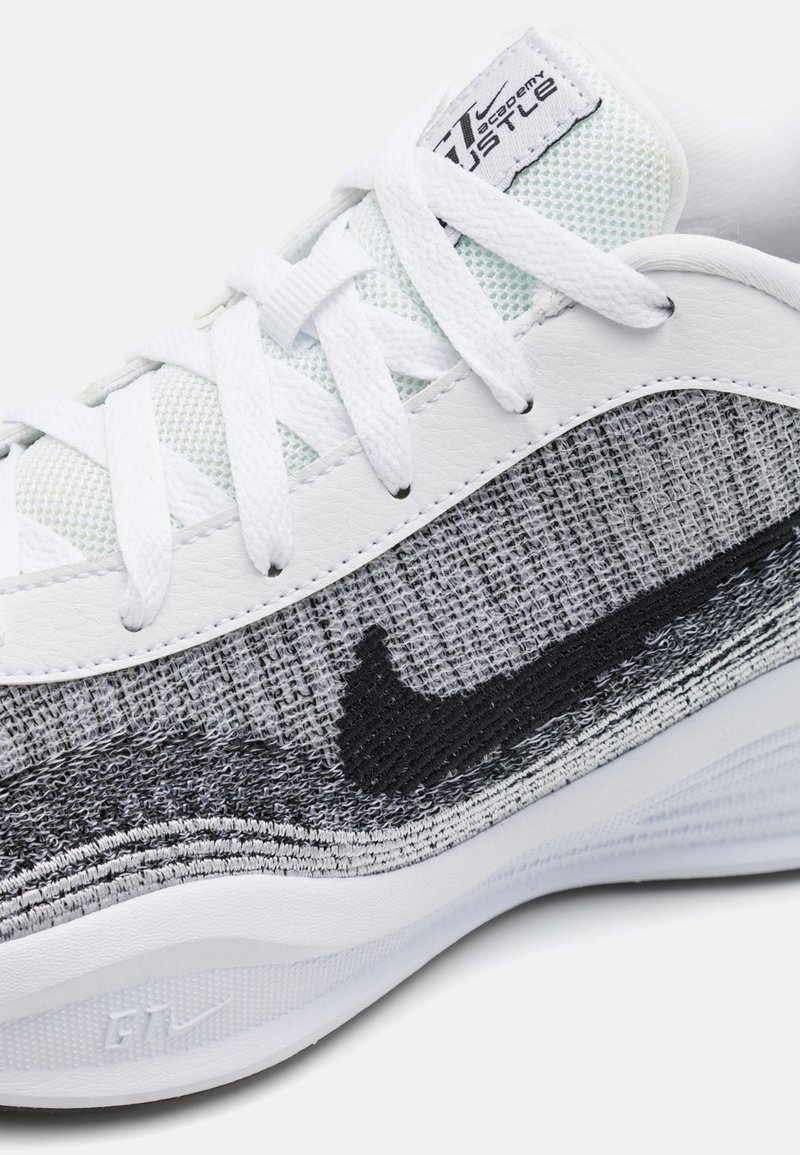 Baskets de sport blanches et grises avec tige en textile, détails en mesh, logo swoosh noir et accents en tissu texturé. Semelle plate en caoutchouc.