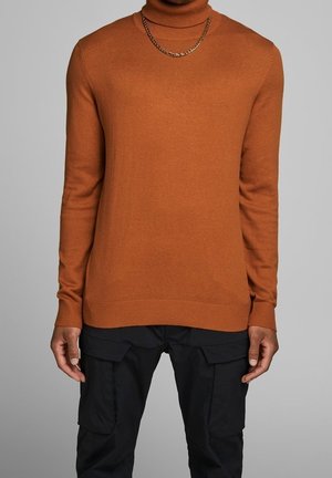Homme portant un pull col roulé orange rouille, une chaîne en or, et un pantalon cargo noir devant un fond gris uni.