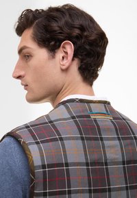 Kostkovaná vesta v šedé, černé, červené a žluté barvě s texturovaným povrchem. Na zadní straně se nachází barevně různorodý textilní akcent.