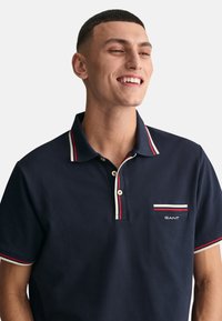 Polo navy con cuello y dobladillos de mangas a rayas en rojo y blanco. Cuenta con un pequeño bolsillo en el pecho con una marca en texto blanco.