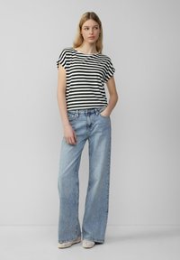 Gestreiftes schwarz-weißes Baumwoll-T-Shirt mit kurzen Ärmeln, kombiniert mit hellblauen weit geschnittenen Jeans. Das Modell steht mit einer Hand in der Tasche.