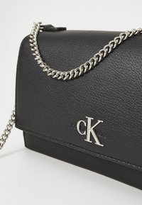 Borsa a tracolla nera in similpelle con manico a catena argentata e logo "CK" in rilievo. Presenta una superficie liscia e una forma rettangolare.