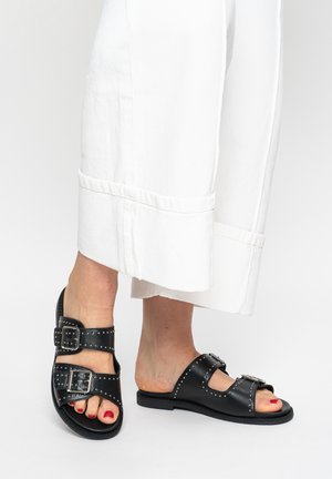 Feet wearing black studded leather slide sandals with buckles, paired with white wide-leg cropped pants.
