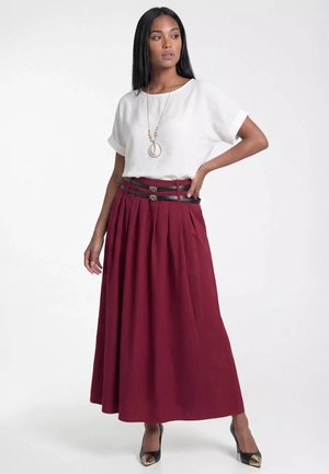 Kobieta w białym topie z krótkim rękawem, długiej bordowej plisowanej spódnicy z podwójnym czarnym paskiem, czarnych szpilkach i naszyjniku z zawieszką, pozuje z ręką na biodrze.