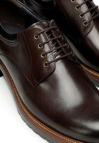 Brązowe skórzane buty mają gładką fakturę, zaokrąglony nosek oraz sześć dziurek na sznurowadła z metalowymi, złotymi okuciami. Podeszwa posiada kontrastujące przeszycia.