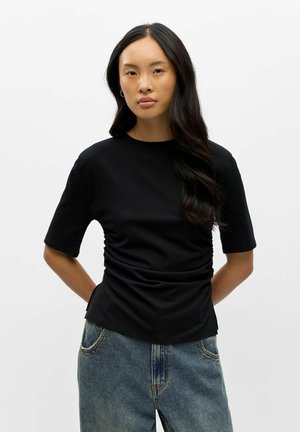 Jeune femme aux longs cheveux foncés portant un haut noir froncé à manches courtes et un jean bleu, debout devant un fond blanc.