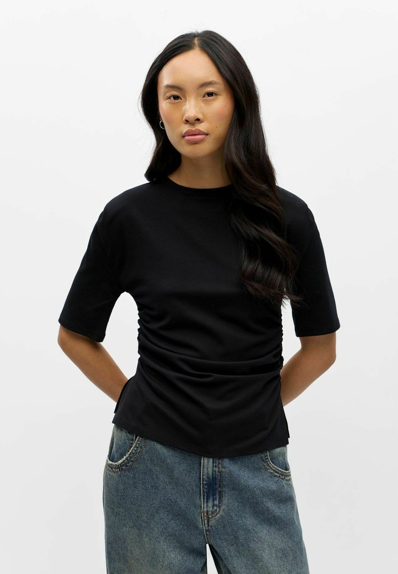Jeune femme aux longs cheveux foncés portant un haut noir froncé à manches courtes et un jean bleu, debout devant un fond blanc.
