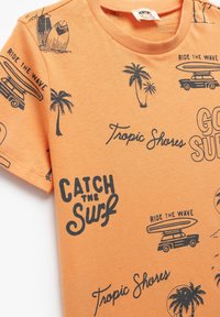 Oranje katoenen t-shirt met zwarte bedrukte graphics, waaronder surfplanken, palmbomen en de teksten "Catch the Surf" en "Ride the Wave."