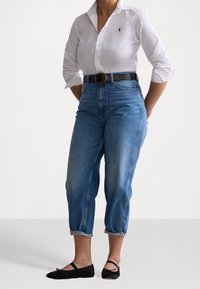 Jeans de mezclilla azul claro de cintura alta con un corte relajado, con los puños enrollados, combinados con una camisa blanca de manga larga y unos zapatos de ballet negros.