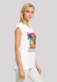 White cotton t-shirt with short sleeves, featuring a colorful graphic design of a tiger, palm trees, and text, paired with light-colored pants.