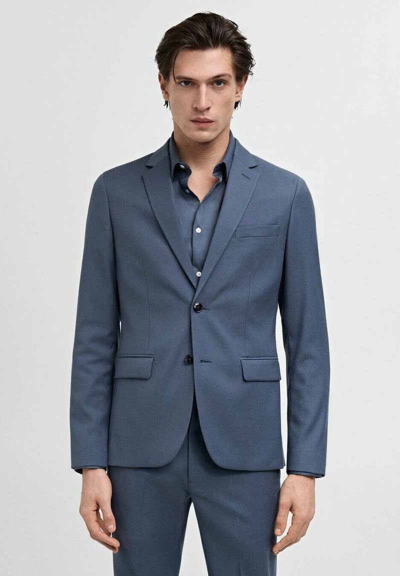 Jachetă de costum bleumarin, ajustată, cu rever în formă de V, două buzunare frontale și închidere cu un singur nasture, asociată cu o cămașă elegantă asortată.