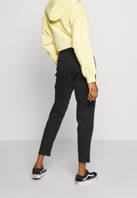 Svarta högmidjade jeans med avsmalnande ben; kombinerade med en gul huhoodtröja. Modellen bär svarta sneakers med vita detaljer.