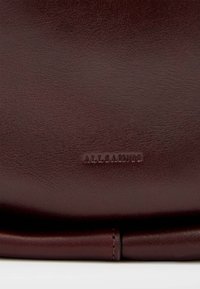 Bolsa de mão de couro bordeaux escuro com uma textura suave e logotipo em relevo "ALLSAINTS". Apresenta linhas limpas e um design minimalista.