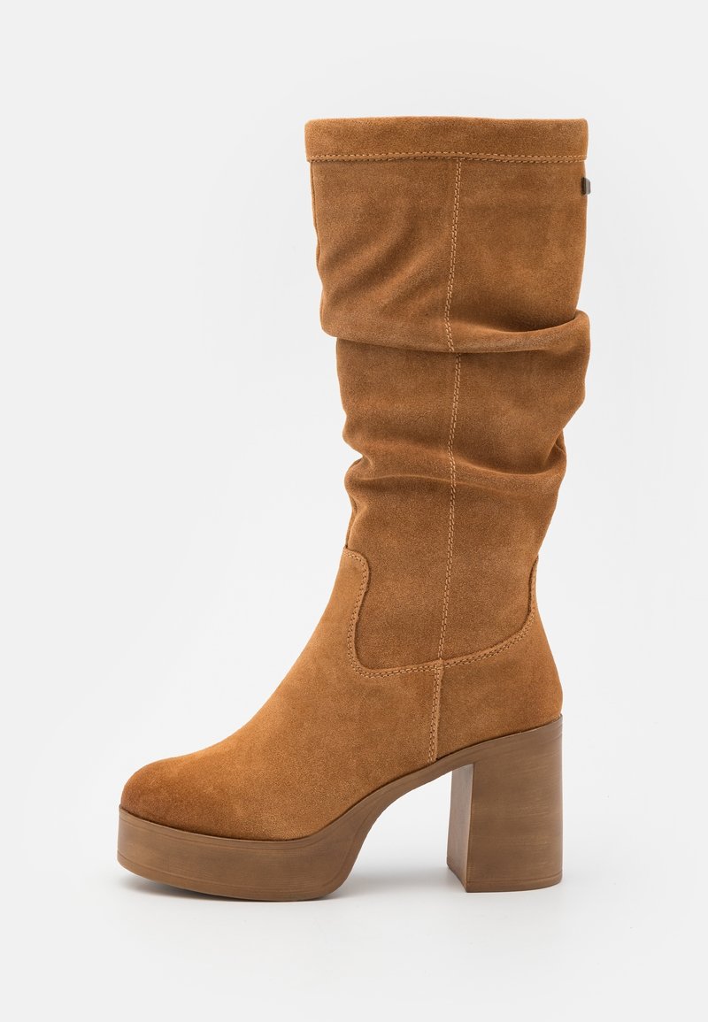 Botte haute en daim marron avec un design souple, un bout arrondi et un talon carré épais. Présente des détails de surpiqûres et une texture lisse.