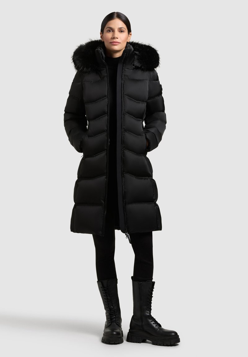Sort pustejakke med hætte med pelskant, der har en lang silhuet, quiltet tekstur, en frontlynlås og lommer. Bæres med sorte leggings og støvler.