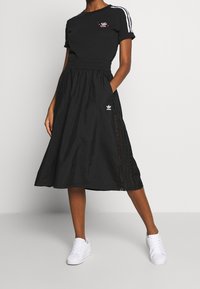 Svart bomull t-shirt med tre vita ränder på axlarna och en liten grafisk design. Svart kjol med elastisk midja och spetsdetaljer i sidorna. Vita sneakers.