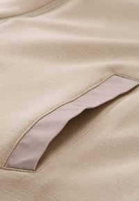 Beige stof met een gladde textuur; heeft een diagonale zakopening met een contrasterende lichtbeige rand langs de randen.