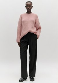 Hellrosa Strickpullover mit lockerer Passform, kombiniert mit hochtaillierten schwarzen Hosen. Das Model steht vor einem neutralen Hintergrund. Schwarze Slipper runden das Outfit ab.