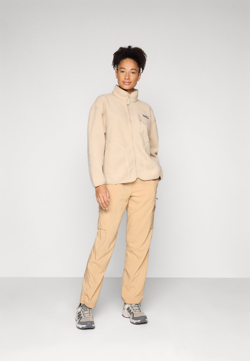 Frau trägt beige Fleecejacke, braune Cargohose und graue Wanderschuhe und steht vor einem einfarbigen hellgrauen Hintergrund.